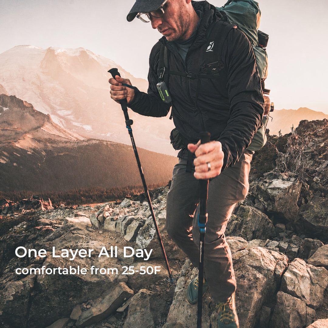 A man hiking on rocky mountains wearing a black one-layer jacket, brown pants, and a green backpack, with text overlay reading 'One Layer All Day comfortable from 25-50F'.