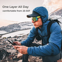 A man in a blue hooded outdoor jacket and reflective sunglasses crouches in a rocky mountain landscape, holding a phone. Text on the left reads 'One Layer All Day' and 'comfortable from 25-50F'.