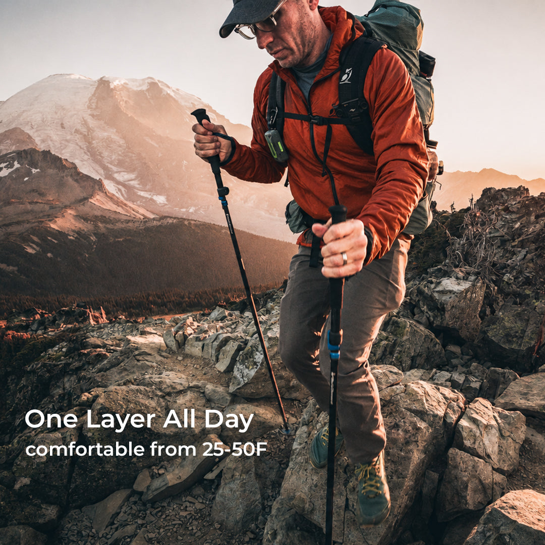 A man hiking on rocky mountainside wearing a one-layer jacket, brown pants, and a green backpack, holding trekking poles. Text overlay reads 'One Layer All Day comfortable from 25-50F'.