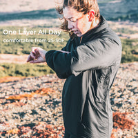 A woman outdoors wearing a dark gray one-layer puffer jacket, pulling her sleeves in to demonstrate the layering system. Text on the left reads 'One Layer All Day' and 'comfortable from 25-50F'.