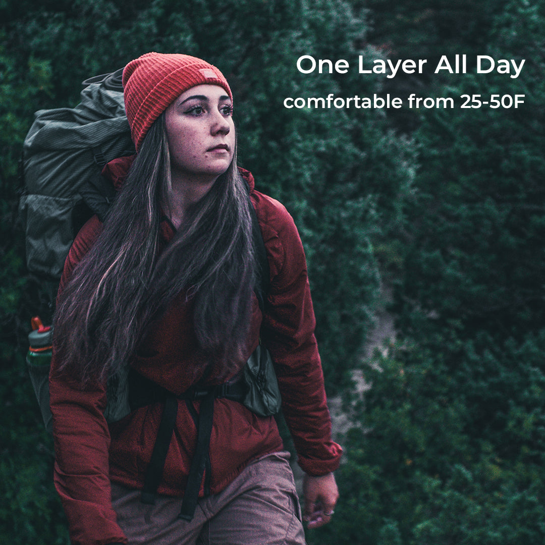 A woman hiking outdoors wearing a rust red one-layer jacket, a red knit beanie, and carrying a large dark green backpack. White text on the right reads 'One Layer All Day' and 'comfortable from 25-50F'.