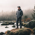 A man wearing a black quilted puffer jacket, dark gray pants, and a black cap stands on a mossy rock in a misty outdoor river setting with trees in the background.