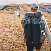 Rear view of a hiker carrying the Outdoor Vitals Shadowlight backpack loaded with gear in mesh front and side pockets, trekking across an open alpine landscape.