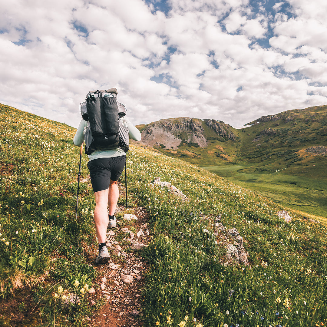 Hiker wearing the Outdoor Vitals Shadowlight backpack, trekking uphill on a narrow mountain trail surrounded by green hills and wildflowers.