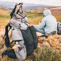 Rear view of Outdoor Vitals Shadowlight backpack on the ground while hiker wearing a jacket sits down on an open alpine landscape.