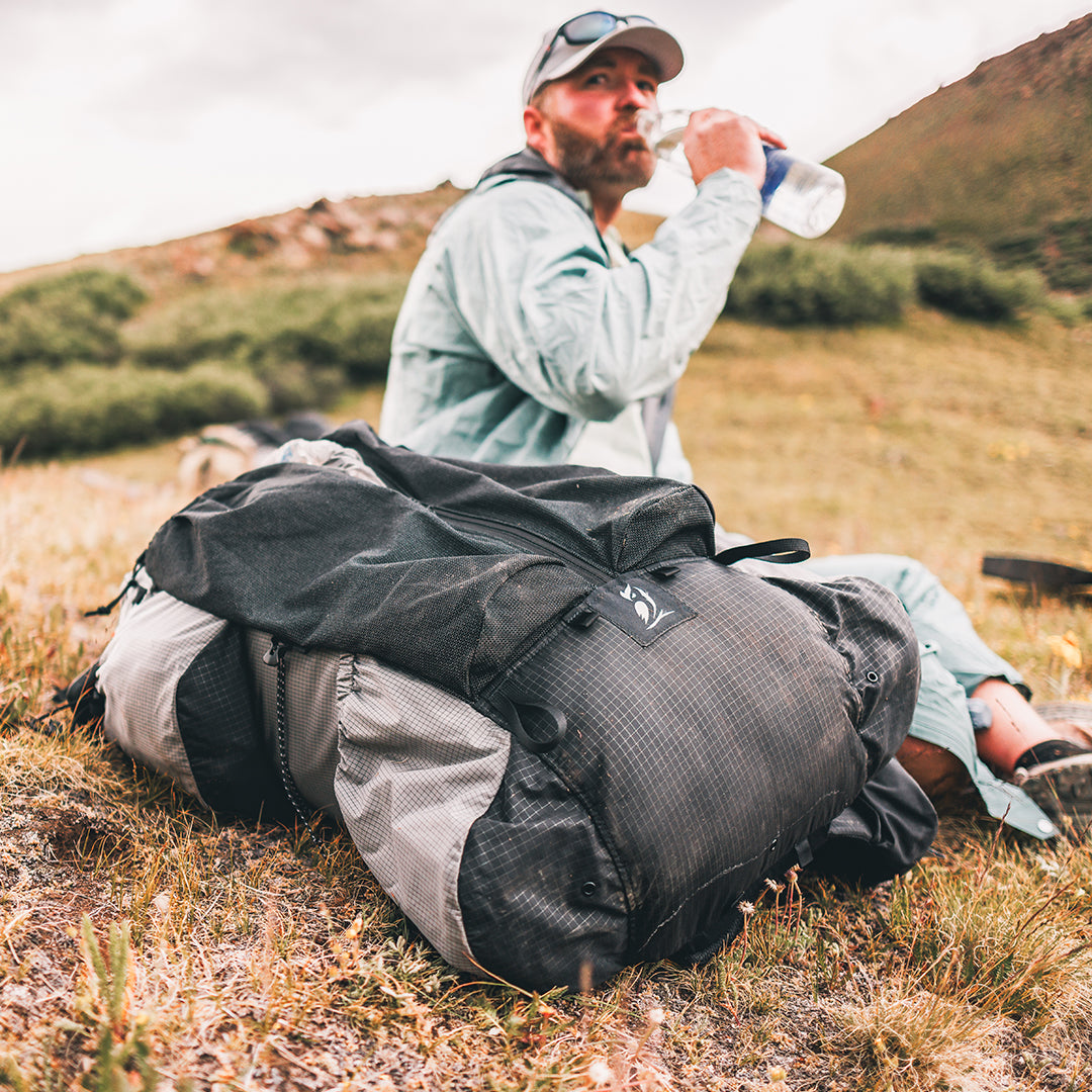 Outdoor Vitals Shadowlight backpack lying on the grass while a hiker drinks water in the background during a trail break.
