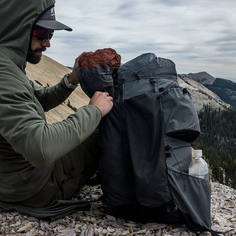 Backpacker in a green jacket loading gear into the front mesh pocket of an Outdoor Vitals Shadowlight backpack while sitting on rocky terrain with forested mountains in the background.