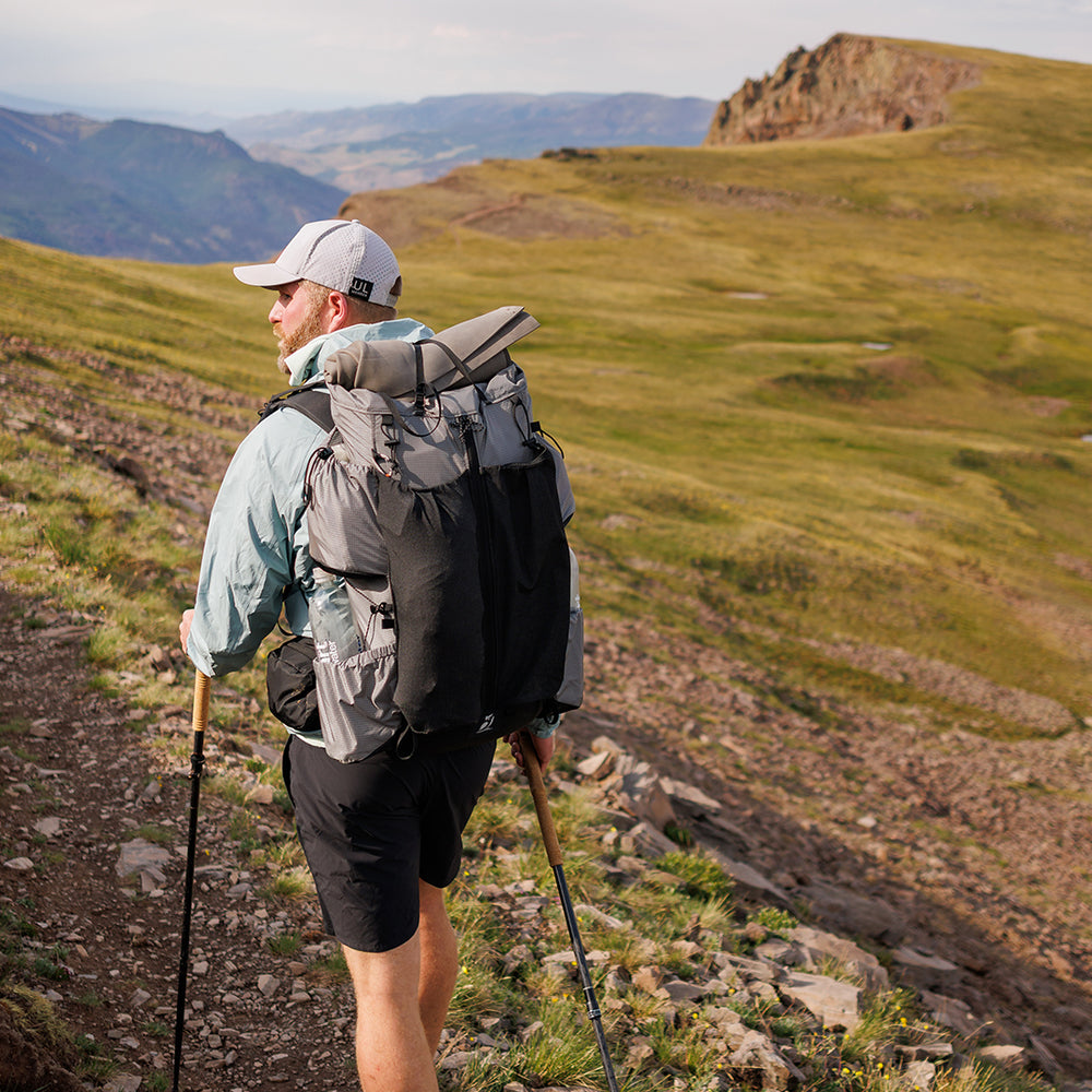 Back view of a hiker in shorts wearing the Outdoor Vitals Shadowlight backpack with a sleeping pad attached, ascending a sunlit alpine ridge.