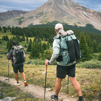 Two hikers on a mountain trail with a large rocky peak and evergreen forest in the background. The hiker in the foreground wears a jacket, shorts, and a large backpack, using trekking poles.