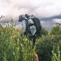 A person hiking through tall green brush under a cloudy sky, wearing a hooded jacket and carrying a large backpack with a sleeping pad and gear.