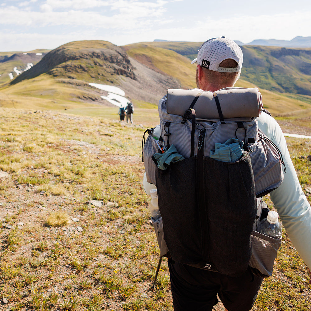 Rear view of a hiker carrying the Outdoor Vitals Shadowlight backpack loaded with gear in mesh front and side pockets, trekking across an open alpine landscape.