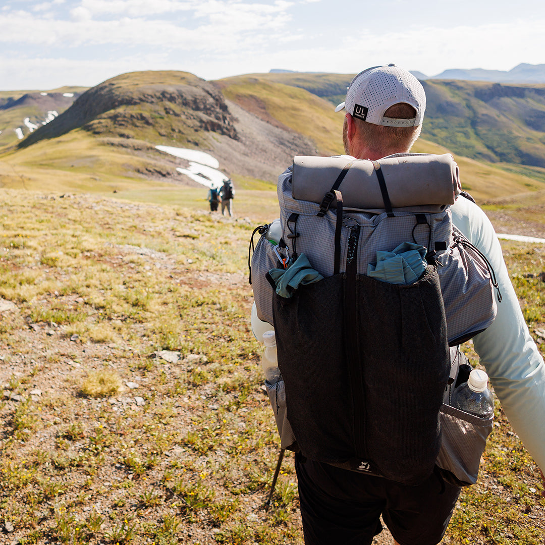 Rear view of a hiker carrying the Outdoor Vitals Shadowlight backpack loaded with gear in mesh front and side pockets, trekking across an open alpine landscape.
