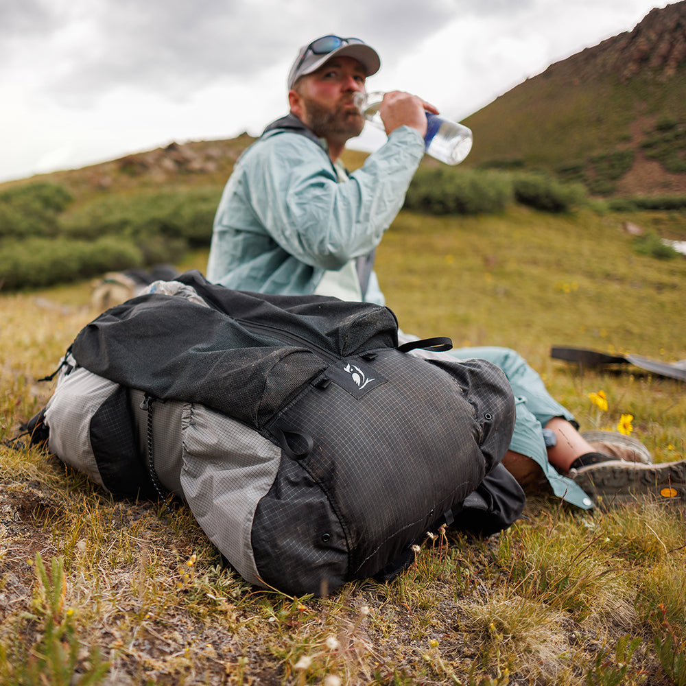 Outdoor Vitals Shadowlight backpack lying on the grass while a hiker drinks water in the background during a trail break.