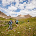 Three hikers carrying Outdoor Vitals Shadowlight backpacks trekking uphill on a trail through a green alpine meadow with rocky peaks in the background.
