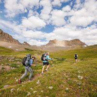 Three hikers carrying Outdoor Vitals Shadowlight backpacks trekking uphill on a trail through a green alpine meadow with rocky peaks in the background.