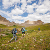 Three hikers carrying Outdoor Vitals Shadowlight backpacks trekking uphill on a trail through a green alpine meadow with rocky peaks in the background.