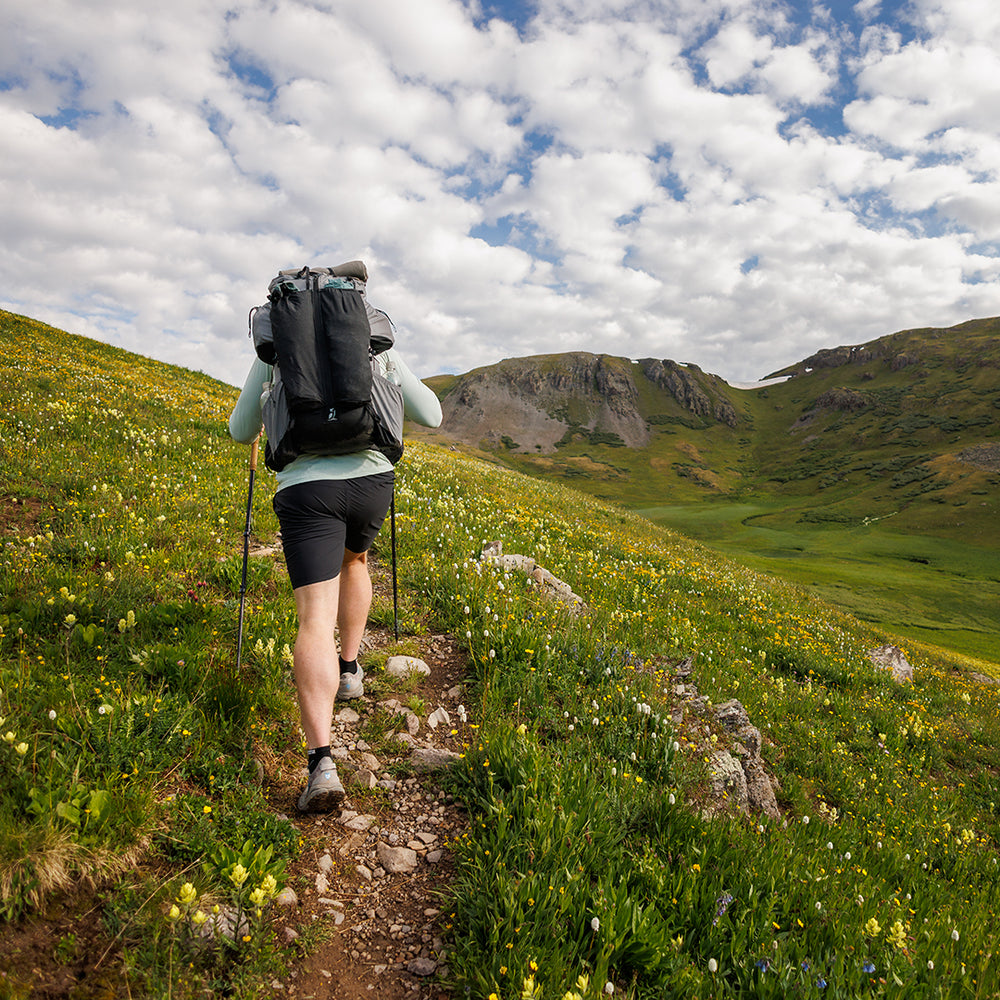Hiker wearing the Outdoor Vitals Shadowlight backpack, trekking uphill on a narrow mountain trail surrounded by green hills and wildflowers.
