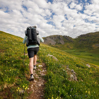 Hiker wearing the Outdoor Vitals Shadowlight backpack, trekking uphill on a narrow mountain trail surrounded by green hills and wildflowers.