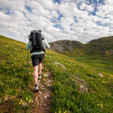 Hiker wearing the Outdoor Vitals Shadowlight backpack, trekking uphill on a narrow mountain trail surrounded by green hills and wildflowers.