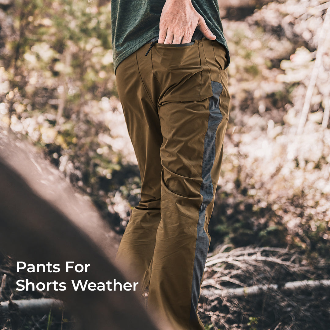 A person outdoors wearing brown pants with a dark gray side panel, shown from the waist down in a wooded setting. White text in the lower left reads 'Pants For Shorts Weather'.