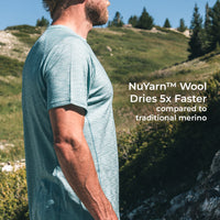 A man wearing a light heathered short-sleeve t-shirt stands outdoors on a grassy mountain slope, with white text on the right reading 'NuYarn™ Wool Dries 5x Faster compared to traditional merino'.