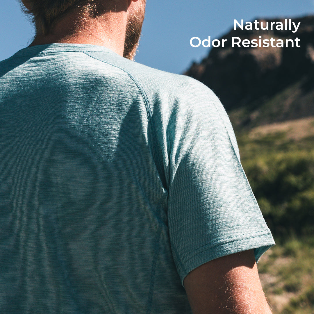 A man wearing a heathered short-sleeve athletic t-shirt, shown from the back in an outdoor mountain setting. White text in the top right corner reads 'Naturally Odor Resistant'.