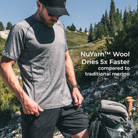 A man wearing a wool short-sleeve athletic t-shirt and black shorts stands outdoors on a rocky mountain path next to a hiking backpack. White text on the right reads 'NuYarn Wool Dries 5x Faster compared to traditional merino'.