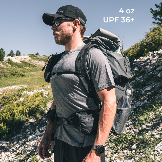A man hiking outdoors wearing a  wool t-shirt with a backpack, set against a rocky, grassy mountain landscape. White text in the upper right reads '4 oz UPF 36+'.
