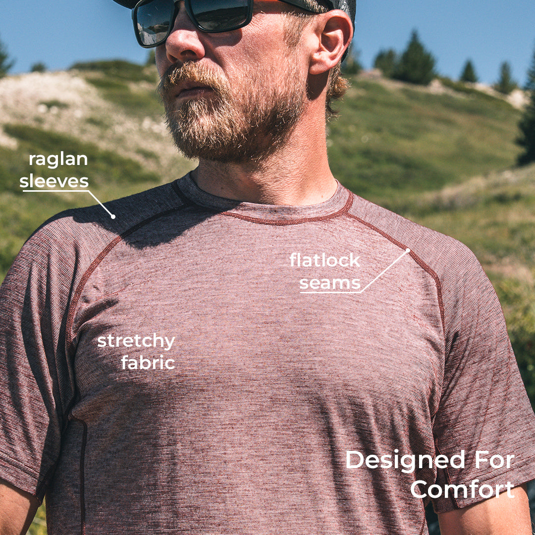 A man wearing a lightweight wool short-sleeve athletic shirt outdoors on a grassy hillside, with white text callouts pointing to raglan sleeves, flatlock seams, stretchy fabric, and 'Designed For Comfort'.