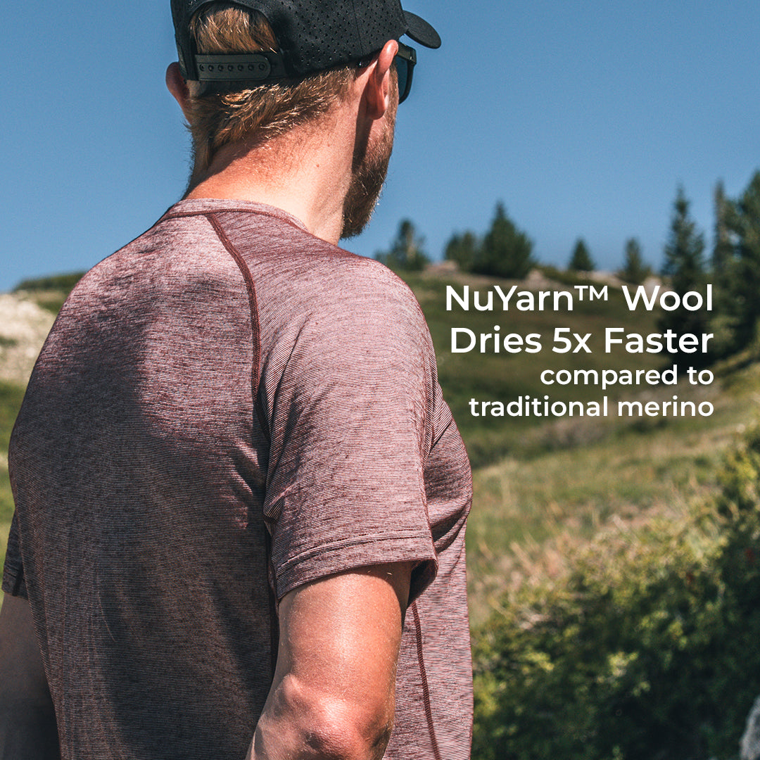 A man wearing a short-sleeve athletic shirt and a black cap, photographed outdoors in a sunny grassy landscape. White text on the right reads: 'NuYarn™ Wool Dries 5x Faster compared to traditional merino'.
