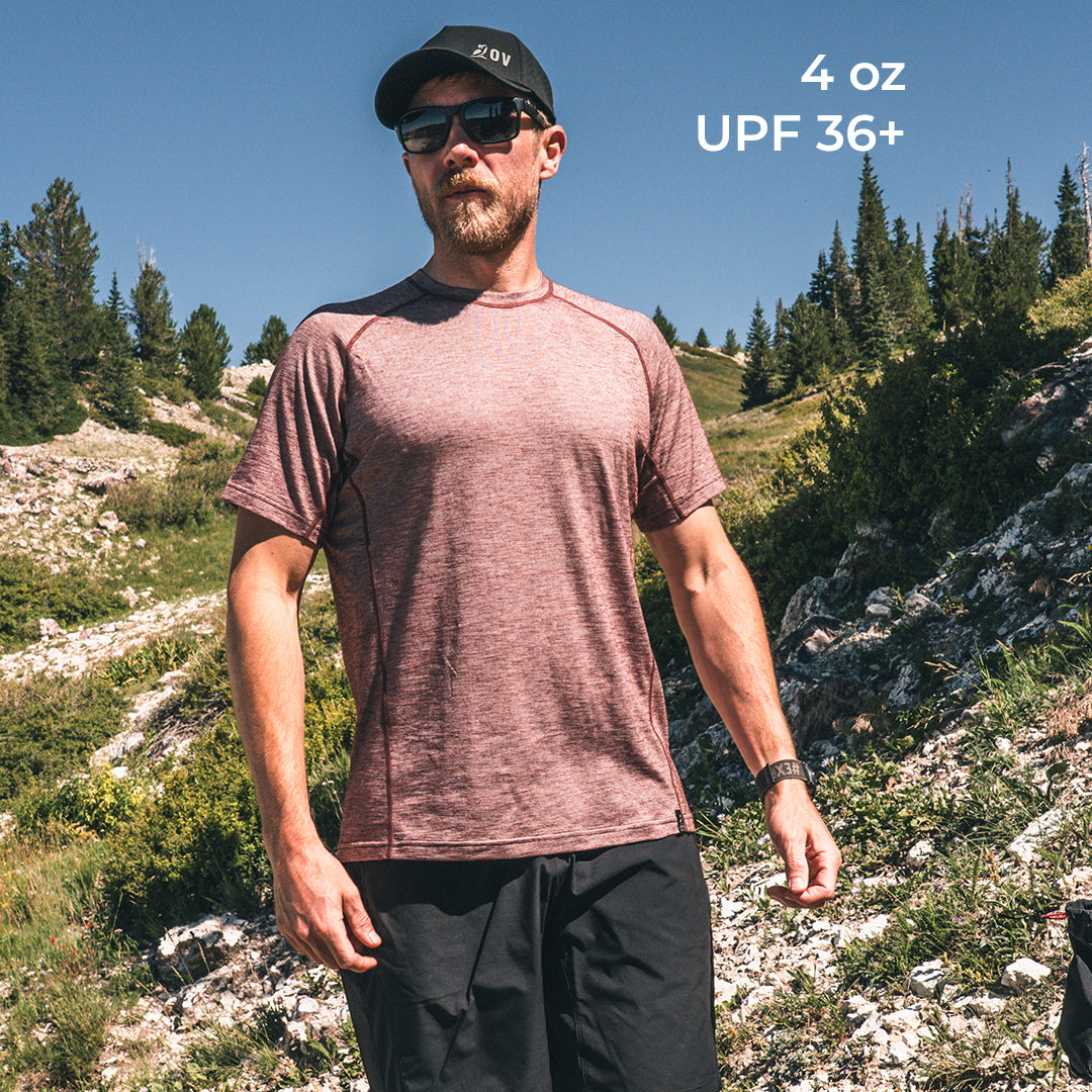 A man wearing a heathered wool athletic t-shirt and black shorts stands outdoors in a rocky, pine-covered mountain landscape under a clear blue sky. The text '4 oz UPF 36+' is visible in the top right corner.