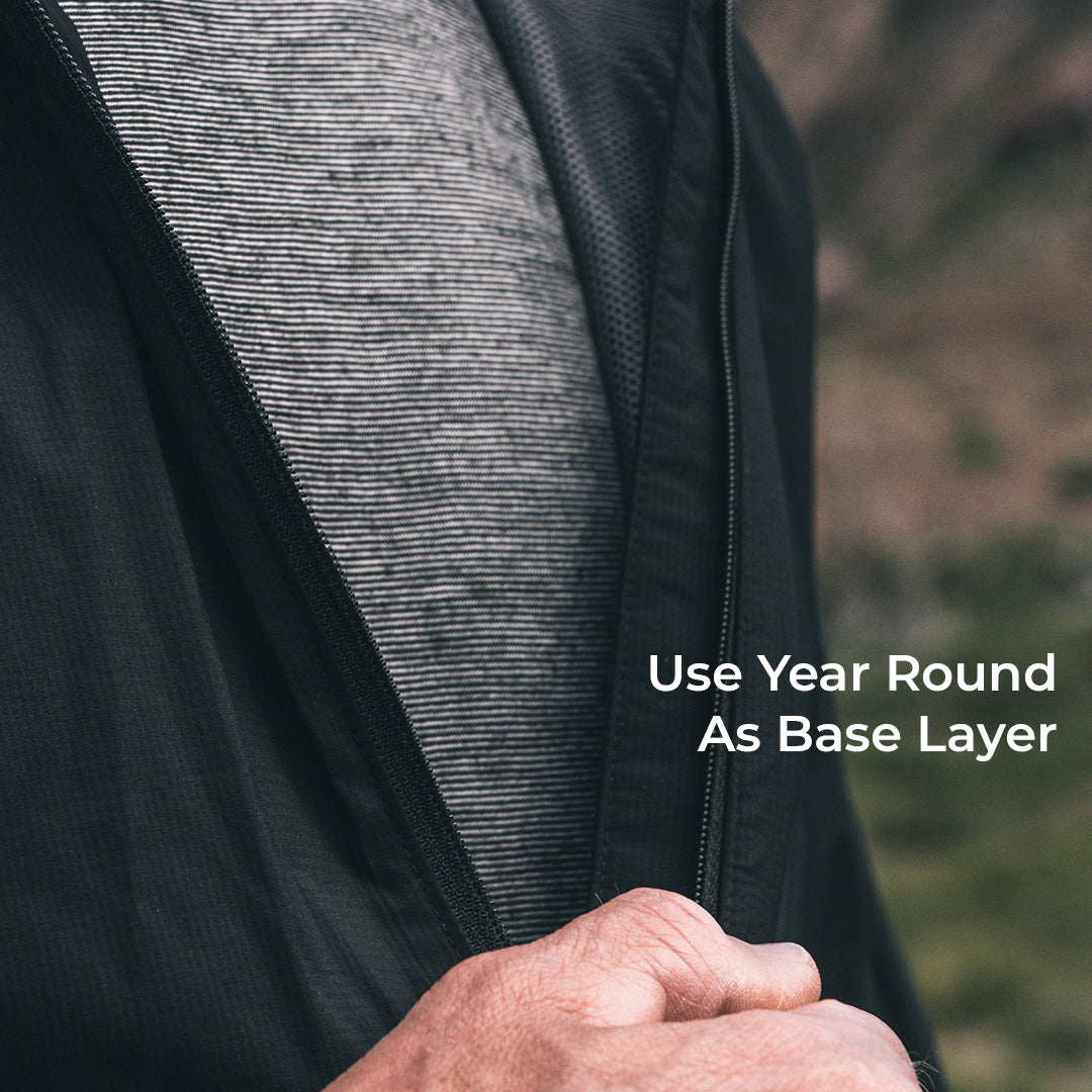 Close-up of a person wearing a black lightweight zip-front layer over a wool base layer, with a blurred outdoor background and the text 'Use Year Round As Base Layer' on the right.