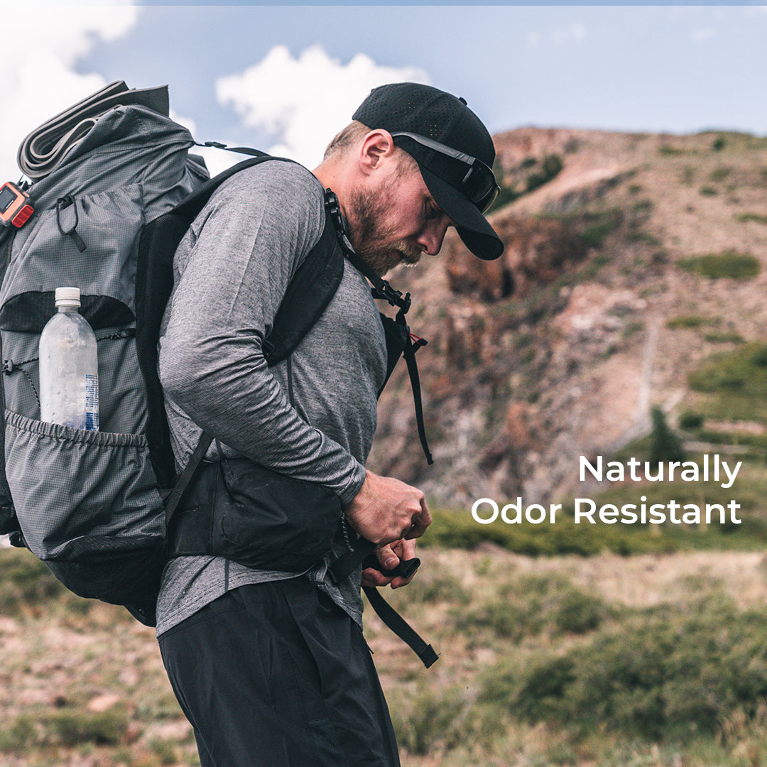A man hiking outdoors wearing a wool sun hoodie, backpack with a water bottle in the side pocket, and a cap. White text on the right reads 'Naturally Odor Resistant'.