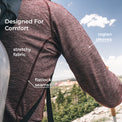 A close-up of a person outdoors wearing a textured merino wool long-sleeve top, with text callouts highlighting stretchy fabric, flatlock seams, and raglan sleeves against a scenic mountain background.