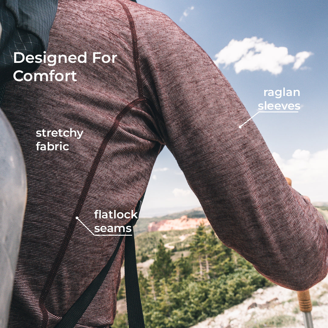 A close-up of a person outdoors wearing a textured merino wool long-sleeve top, with text callouts highlighting stretchy fabric, flatlock seams, and raglan sleeves against a scenic mountain background.