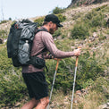 A man hiking on a rocky, grassy mountain slope wearing a large backpack, a long-sleeve wool sun shirt, black shorts, and a black cap while using two trekking poles.