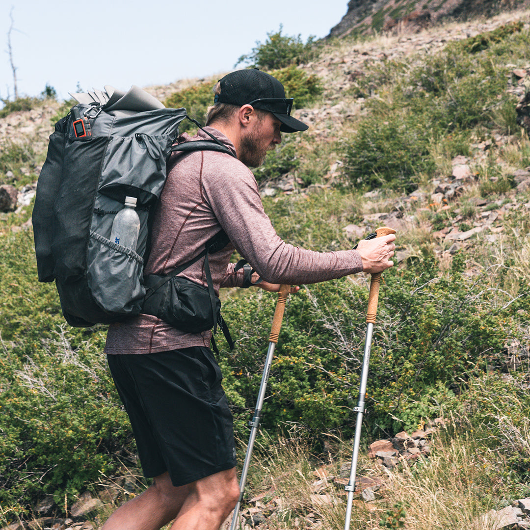 A man hiking on a rocky, grassy mountain slope wearing a large backpack, a long-sleeve wool sun shirt, black shorts, and a black cap while using two trekking poles.