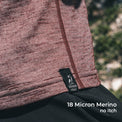 Close-up of a wool sun shirt with a black Outdoor Vitals label, worn outdoors with blurred greenery in the background. White text at the bottom reads '18 Micron Merino no itch'.