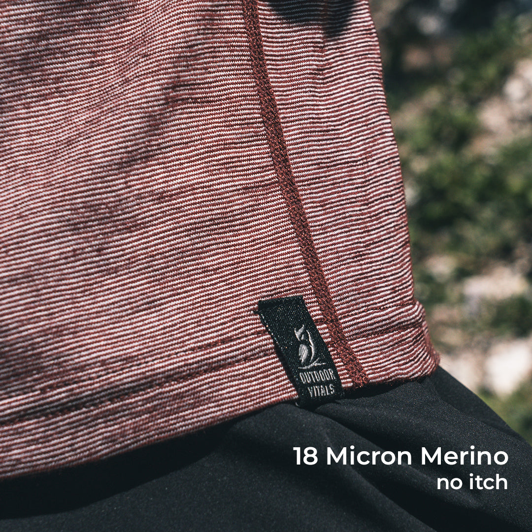 Close-up of a wool sun shirt with a black Outdoor Vitals label, worn outdoors with blurred greenery in the background. White text at the bottom reads '18 Micron Merino no itch'.