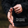A close-up of a person wearing a black thumbloop on their thumb while adjusting a black sleeve, with the text 'Thumbsloops For Layering' on the right side.
