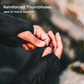Close-up of a person wearing black fingerless gloves and adjusting the thumbhole opening, with overlaid text reading 'Reinforced Thumbholes' and 'seal in more warmth' against a blurred outdoor rocky background.