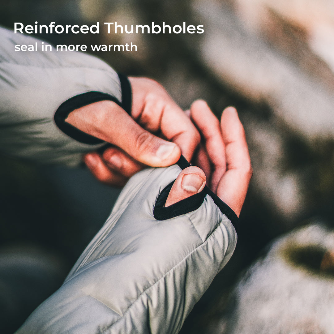 Close-up of a person's hands adjusting a thumbhole on a light gray puffer jacket, with text in the top left reading 'Reinforced Thumbholes' and 'seal in more warmth'.