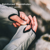 Close-up of a person's hands adjusting a thumbhole on a light gray puffer jacket, with text in the top left reading 'Reinforced Thumbholes' and 'seal in more warmth'.