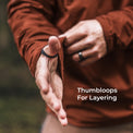 A close-up of a person's hands wearing a black thumbloop on their left thumb, with another black ring on their right hand. The person is wearing a jacket, and the text 'Thumbsloops For Layering' is visible on the bottom right.