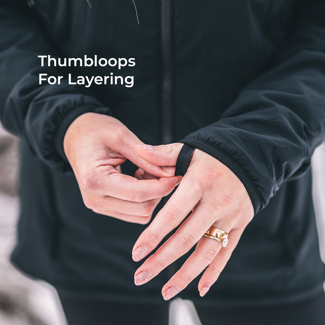 Close-up of a person wearing a black zip-front jacket and fastening a thumbloop on the cuff, with white text reading 'Thumbloops For Layering' on the upper left.