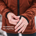 Close-up of a person wearing a rust-brown jacket and fastening a black thumbloop on the cuff of the sleeve. White text on the lower left reads 'Thumbloops For Layering'.
