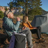 Man and woman wearing insulated down jackets relaxing at a campsite beside a backpacking tent.
