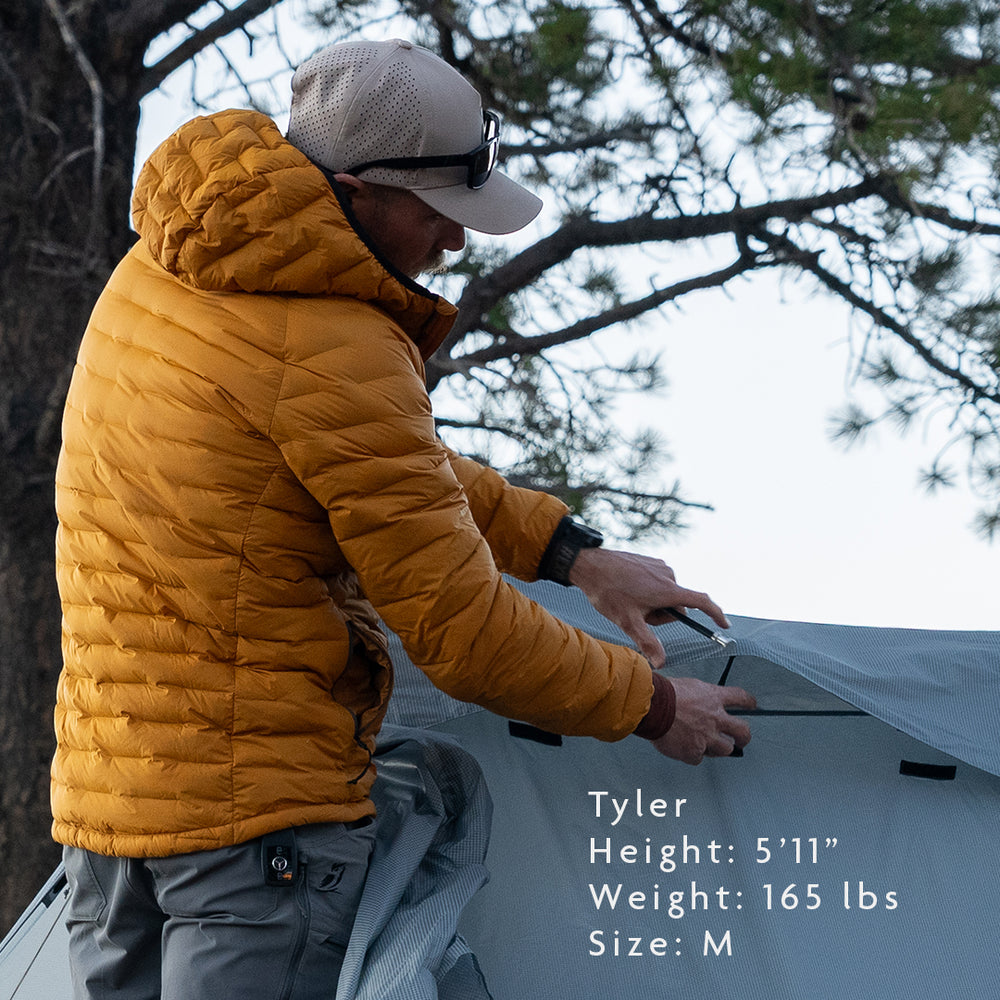 Man wearing insulated hooded down jacket while setting up a tent outdoors; model Tyler is 5'11", 165 lbs, wearing size medium for fit reference.