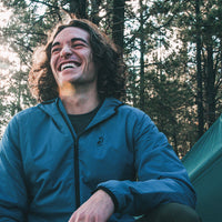A smiling man with curly brown hair wearing a blue zip-up jacket sits outdoors in a forest setting with trees and a tent visible in the background.