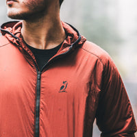 A close-up of a man wearing a hooded zip-up jacket with a small black bird logo on the left chest, photographed outdoors in soft, blurred light.